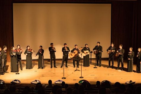 Dressed in black mariachi attire and holding various instruments, a group performs on stage.