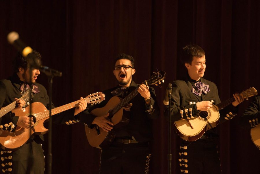 In traditional mariachi attire, someone plays the guitar and sings. Two people with other guitar-like instruments play on either side of him.