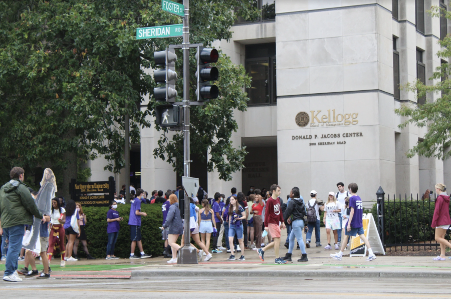 A tan building is in the background as students stand in line in front of it. Other students cross the street at the walk signal.