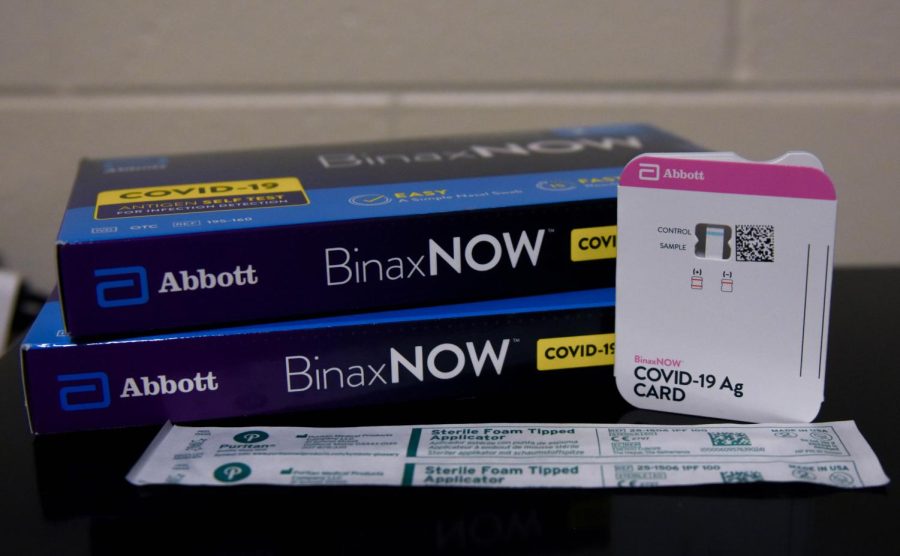 Two blue BinaxNOW rapid antigen test boxes stacked on one another on a black surface. A wrapped nasal swab is underneath with a testing card propped up on the left.