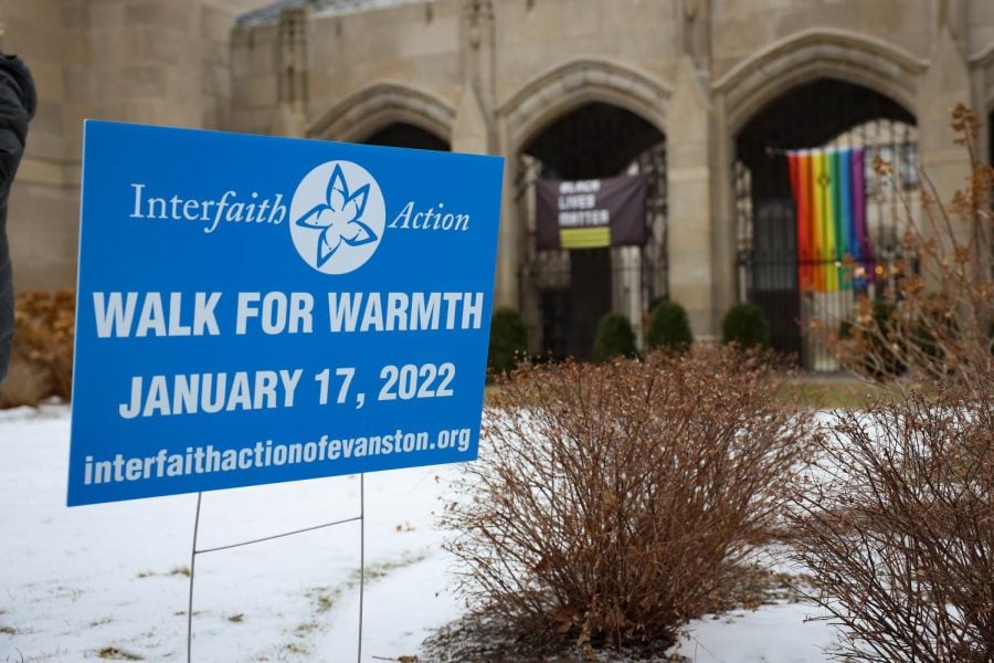 A blue sign reads “Walk for Warmth” in white letters and sits on a snowy ground. A rainbow flag is visible in the background.