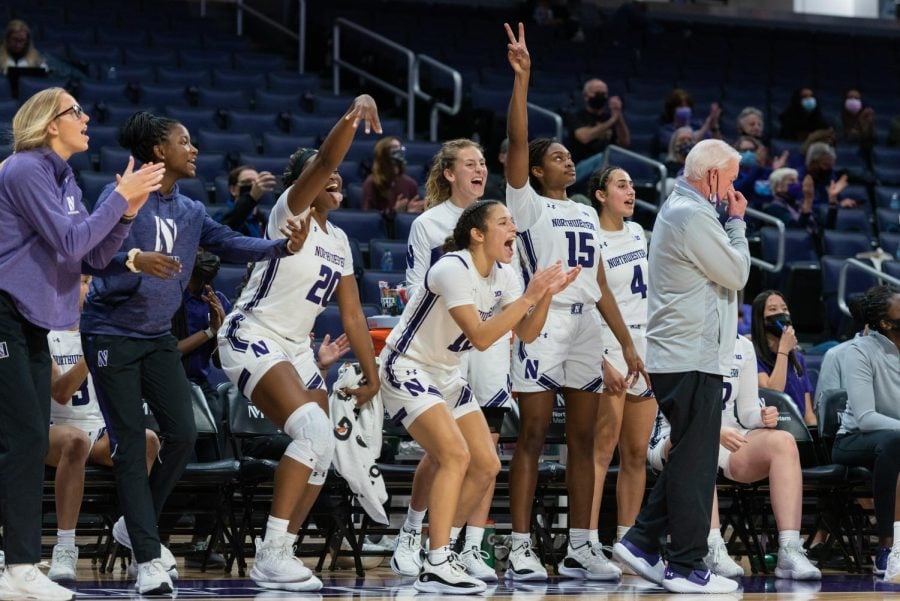 Seven+girls%2C+some+in+white+basketball+jerseys+with+a+purple+stripe+and+others+in+purple+sweatshirts%2C+are+happy+and+excited+from+the+bench.