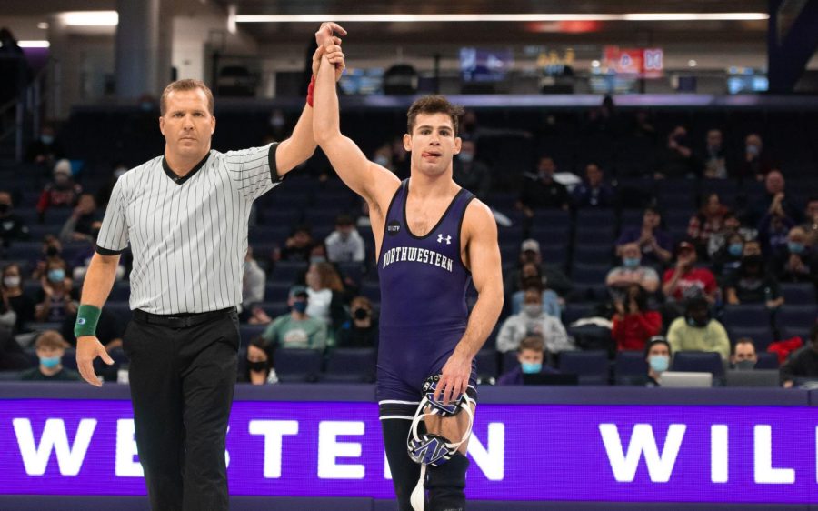 Referee wearing black and white striped shirt holds up the hand of man in a purple singlet who sticks his tongue out.