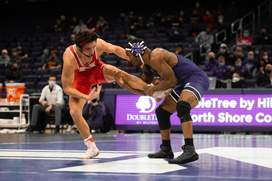Man in purple singlet lifts the leg of a man in red singlet while they are both standing.