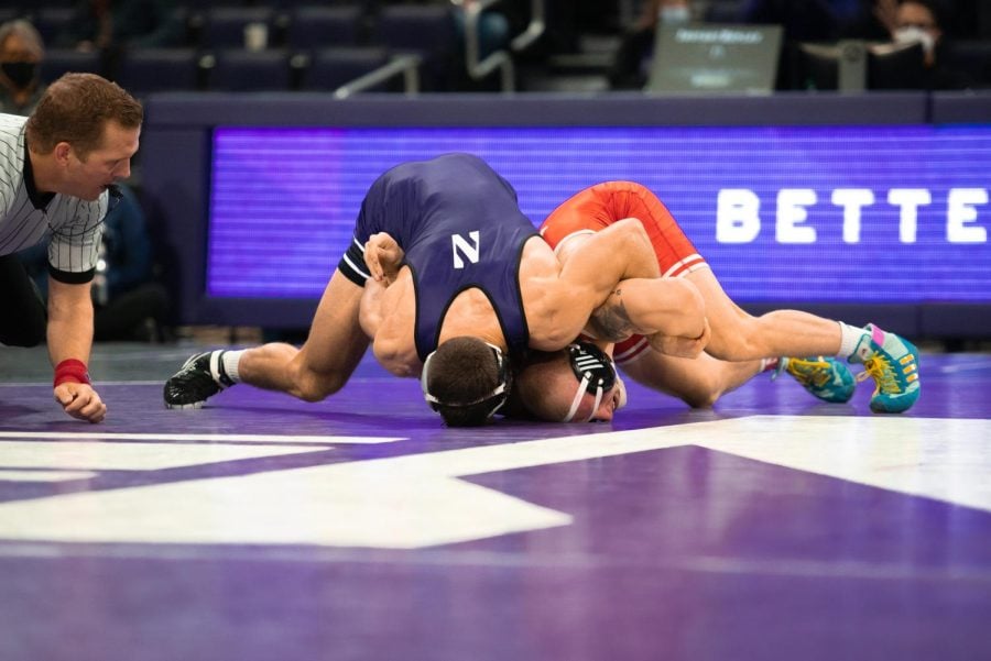 Man in purple singlet wraps arms around the neck of a man in red singlet while they both lay on a wrestling mat. 