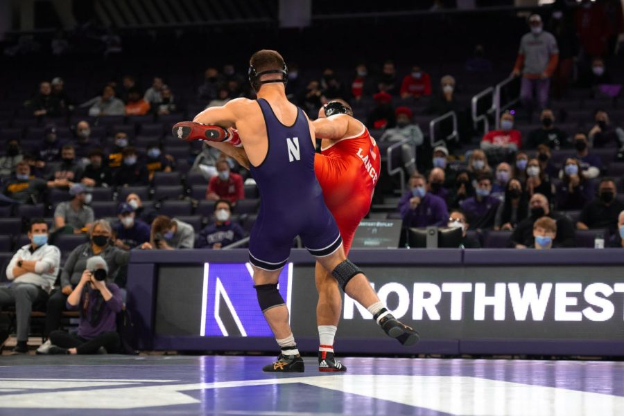 Man in purple singlet lifts the leg of a man in red singlet while they are both standing.