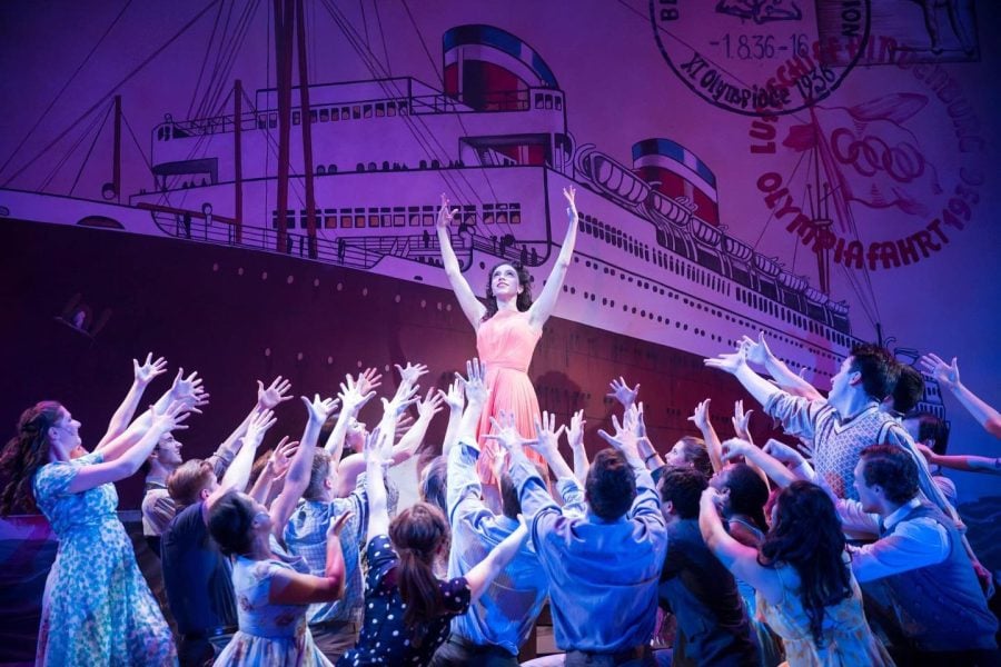A crowd bows with their hands in the air to someone in an orange dress. A spotlight shines from above and a backdrop with a boat is in the background.