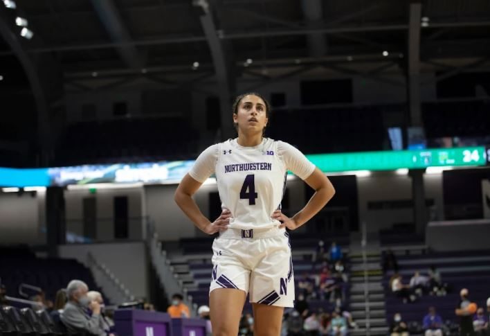 Basketball player wearing white jersey stands with hands on her hips in disappointment.