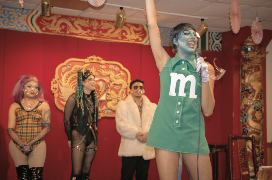 Five performers in various outfits stand behind a performer in a green dress with a microphone on stage.