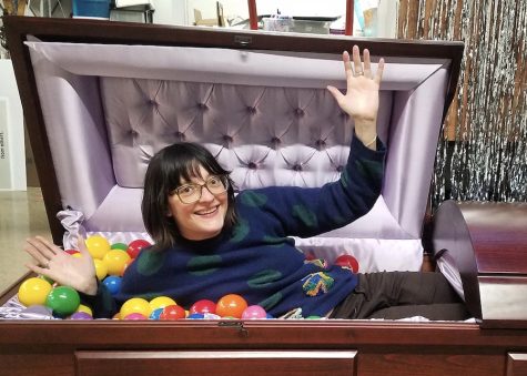 A woman (alive) lies in a casket with her arms up. The casket is filled with colorful plastic balls. 