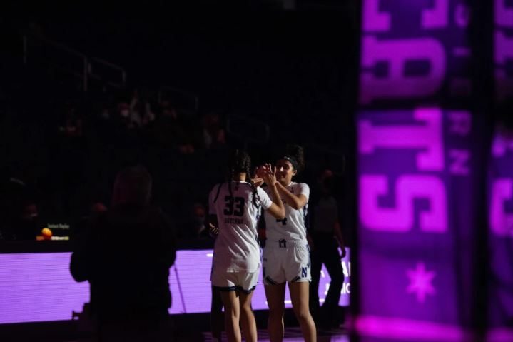 Two girls in white jerseys, one with the number 33 and one with the number 4, high five in a dark stadium, except for a purple light in the foreground.