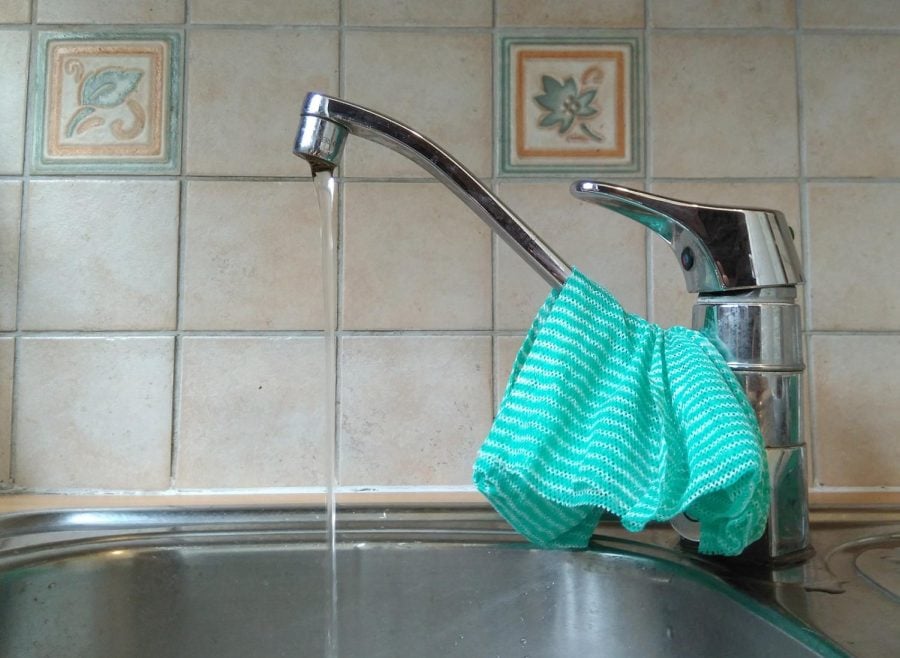 A steel kitchen faucet. The tiles in the back are white, orange and blue. There is a teal towel on the faucet.