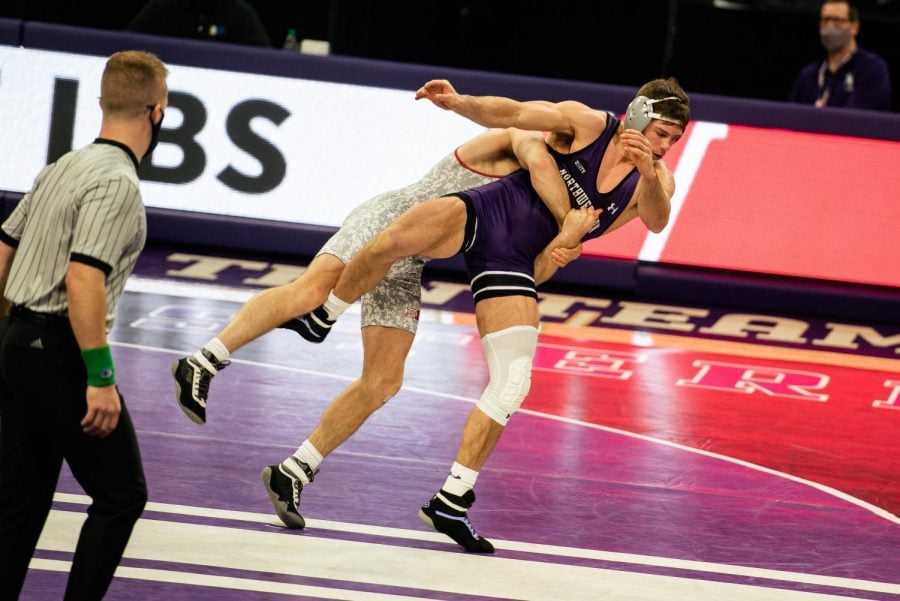 Northwestern wrestler in purple uniform is held across the chest by Wisconsin wrestler in white uniform.