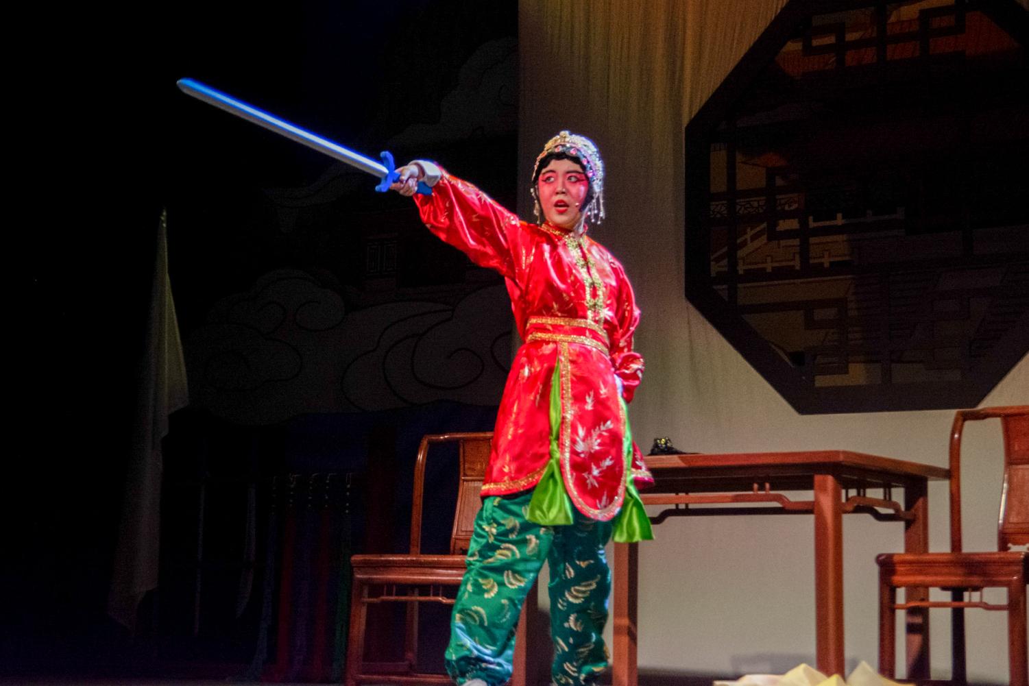 An+actress+dressed+in+red+and+green+traditional+Chinese+dress+stands+onstage+and+points+a+sword+toward+the+audience.