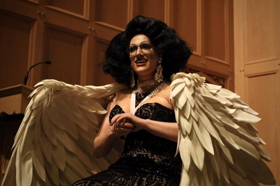 Ms.+Penny+Cost+speaks+to+the+audience%2C+while+wearing+a+black+wig%2C+glasses%2C+crystal+earrings%2C+a+big+necklace%2C+a+black+dress+and+angel+wings.