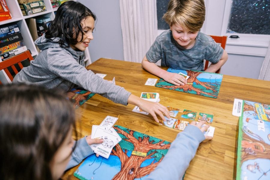 Kids sit around a table. A mat with an illustration of a tree is in front of each, and they’re holding cards. Six illustrated square cards sit in the middle of the table, and one player is reaching towards them.