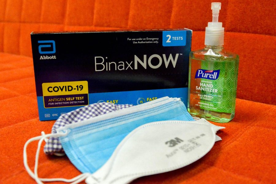 Three types of masks in a pile in front of a black box of COVID-19 tests and a bottle of green hand sanitizer to the right.