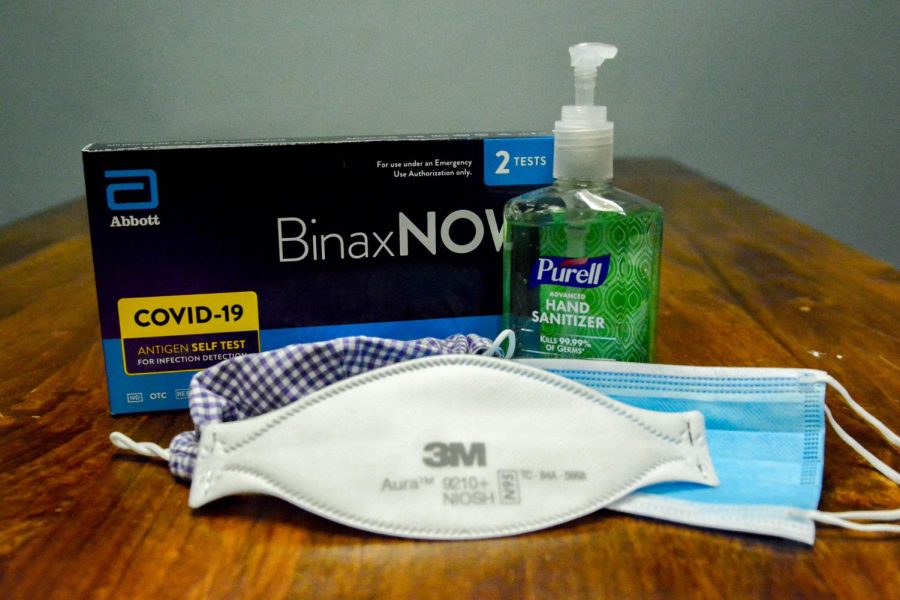 Three types of masks in front of a black box of COVID-19 tests and a bottle of green hand sanitizer.