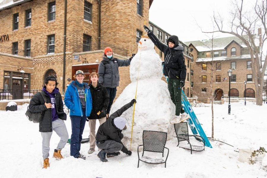 A tall snowman in the fraternity quad with four people standing around it.