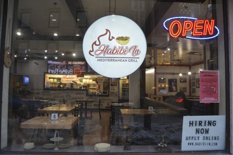 The window looking into Habibi In displays an open sign, vaccination requirements and a “Hiring Now” advertisement. Through the window, there are many tables, a large menu and a man sitting to the right side.