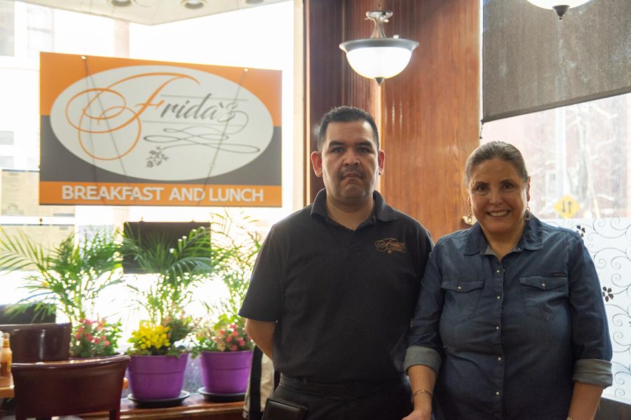Two people pose in front of an orange sign that says “Frida’s” that’s hanging on a window with plants underneath.