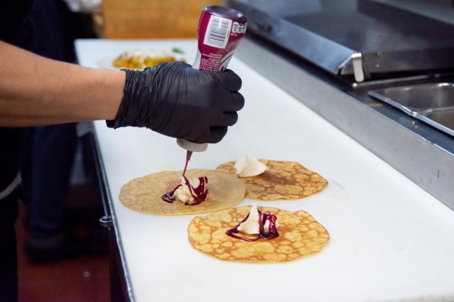 A gloved hand garnishes a crêpe with red strawberry jam. Three crêpes lie on a white table.