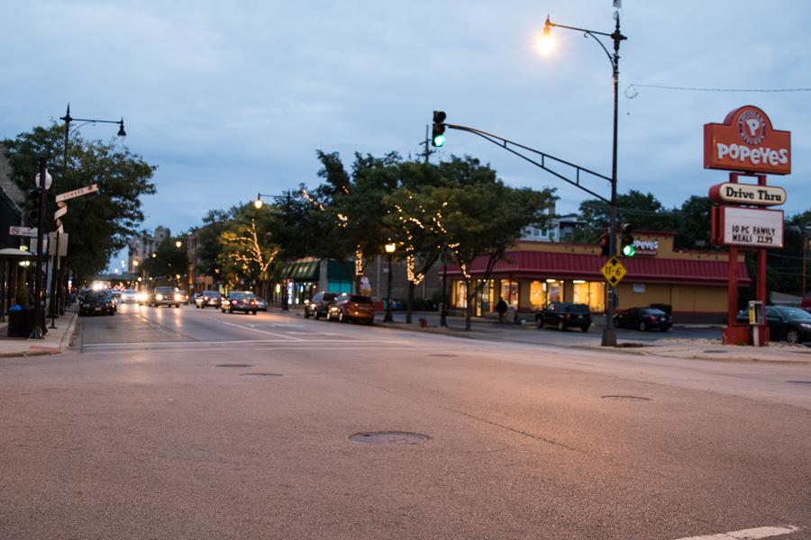 An intersection of Howard Street at dusk. There is a Popeyes at the street corner, the sky is dark, and the streetlights are green and a warm yellow.