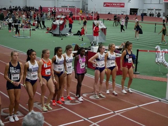 A group of female runners line up before the start of a race on an indoor track.