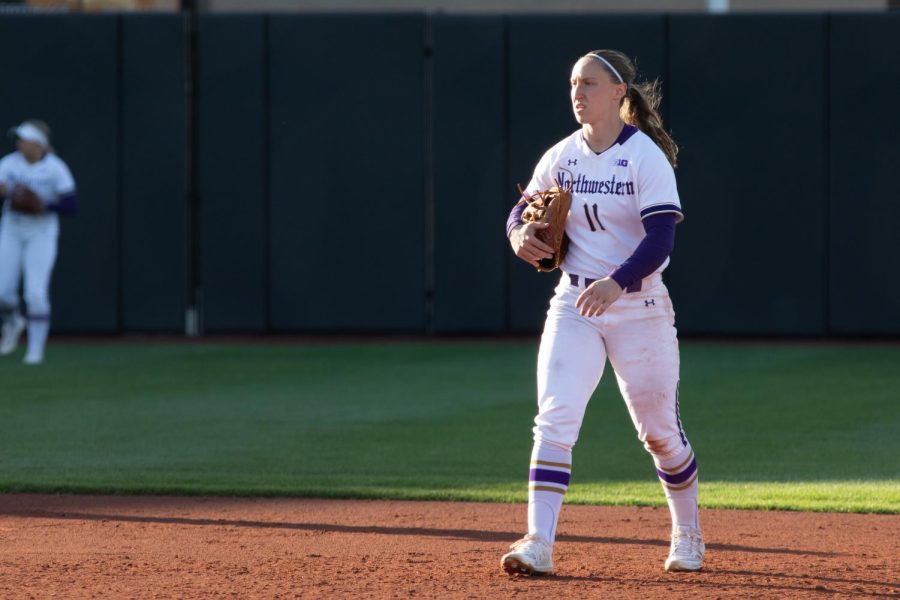 Softball player in white uniform holds glove in hand while standing on infield dirt.