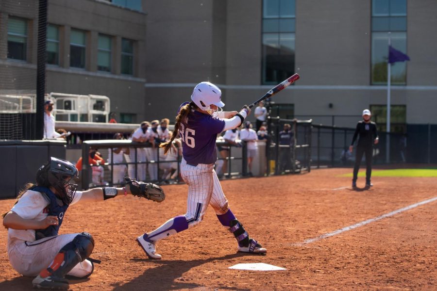 Softball player in purple and white uniform holding a bat swings at a pitch in front of an opposing player wearing catcher’s gear.