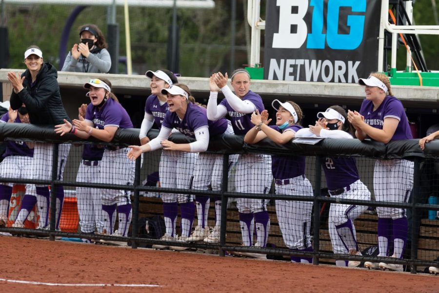 Softball+players+in+purple+and+white+uniforms+climb+the+dugout+wall+and+clap+their+hands.