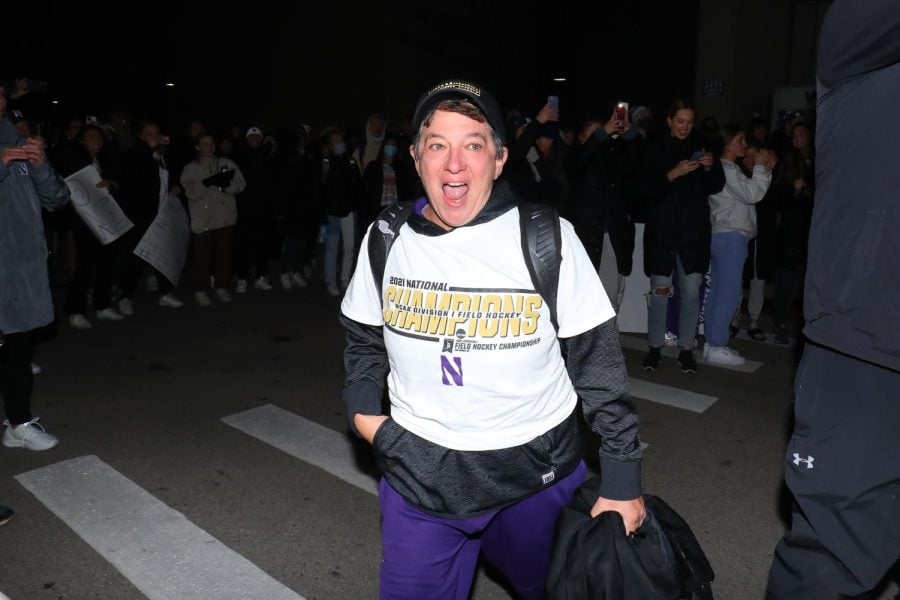 Field Hockey coach in a black hat, white national champions shirt, and purple pants smiles as she walks across a dark street