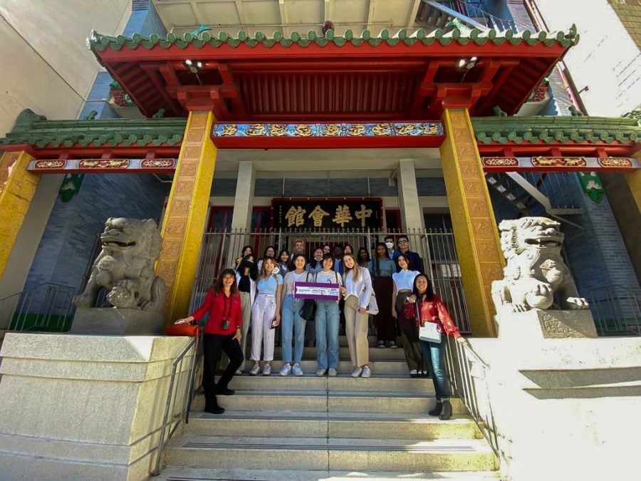 A group of students stand on the steps in between two stone lion statues and under a building with a red and green roof. A sign with gold Chinese characters is above the entrance behind them.