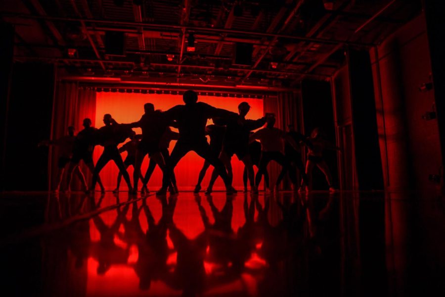 Silhouetted dancers in front of a red backdrop squat at the beginning of their dance.