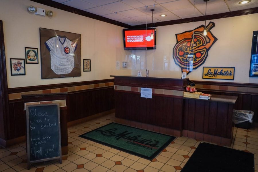 Tiled floor with a green mat with the words “Lou Malnati’s Pizzeria” printed on it in front of a dark brown host stand with clear plexiglass attached. A framed Chicago Cubs jersey and graphic of a pizza with a slice cut out are on the wall.