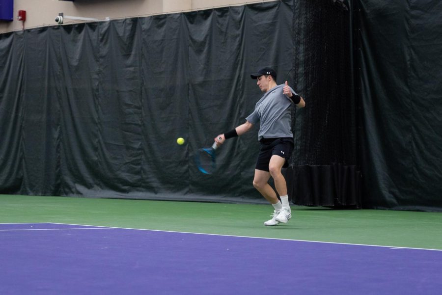 Male tennis player in black hat, gray shirt, black shorts and two black armbands returns shot on purple and green court.