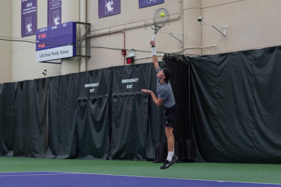 Male tennis player in black hat, gray shirt and black shorts serves the ball on a purple and green court.