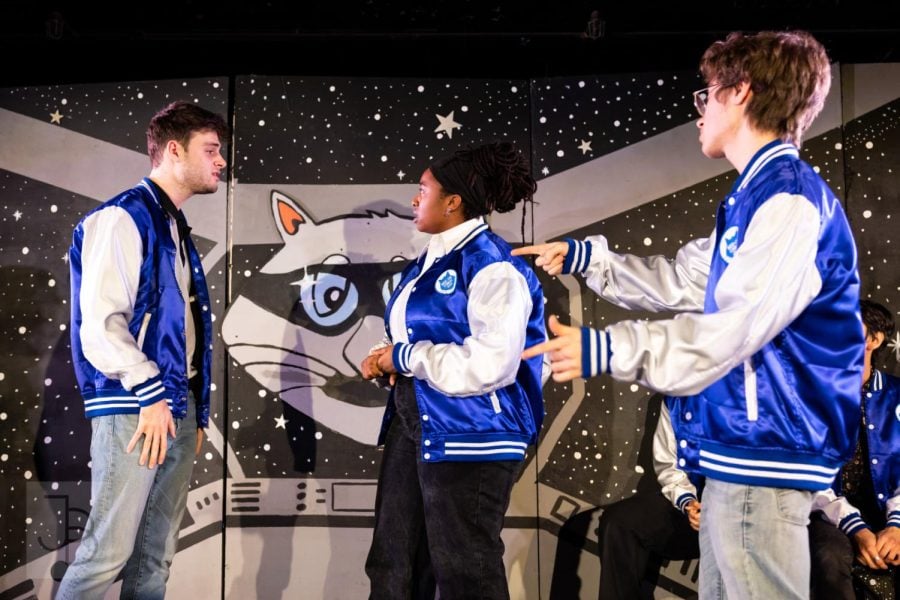 Three+students+wearing+blue+and+white+jackets+are+gesturing+against+a+black+background+showing+stars+and+the+face+of+a+gray+cat.