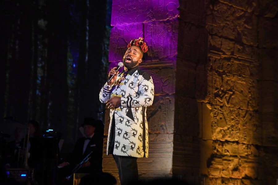 Artist Mykal Kilgore sings wearing a white suit with black patterns. The background lighting is purple and gold.