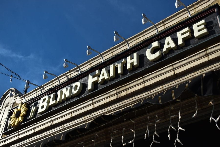 A sign with white letters on a black background reading “Blind Faith Cafe” with a sunflower in front of a blue sky. There are lights on the building around the sign.