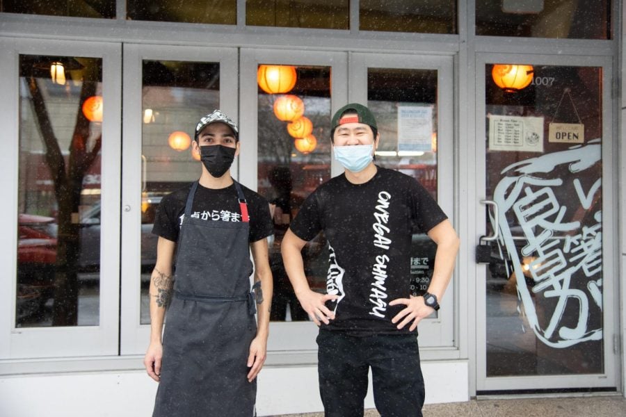 Two employees in black shirts and aprons pose in front of a white storefront.