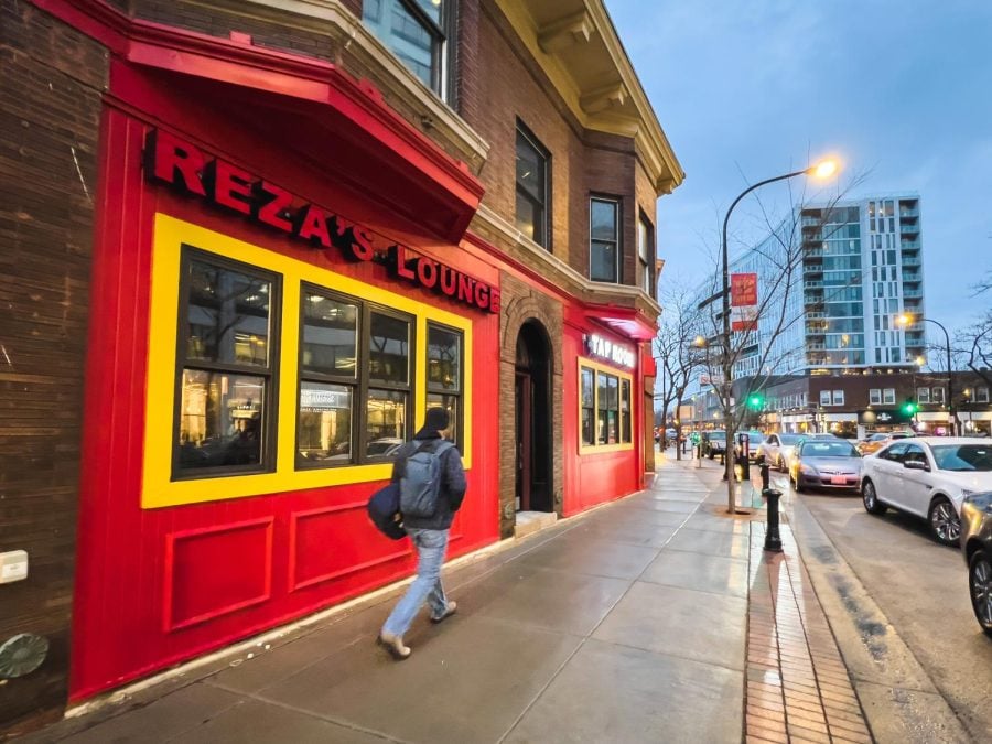 Reza’s Lounge and Tap Room facing the street at dusk.The walls are red and yellow and a person is walking by.