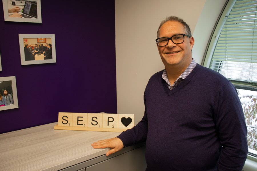 Someone in glasses and a purple sweater smiles in front of a sign that says “SESP” in scrabble tiles.