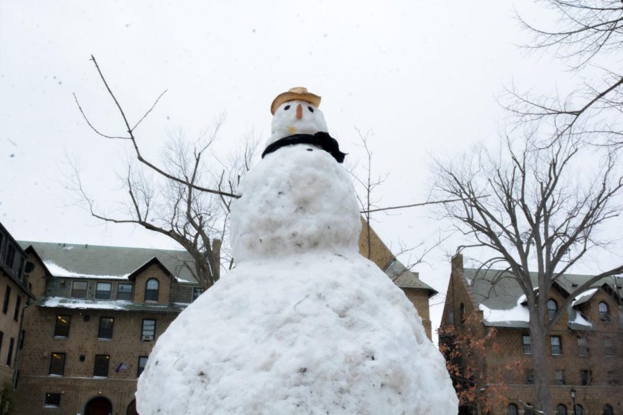 A very large snowman with buildings behind it.