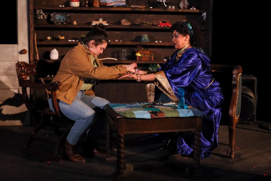 Two actors on stage, sitting across from each other on wooden chairs and holding hands. One wears a blue robe and the other wears jeans and a jacket.