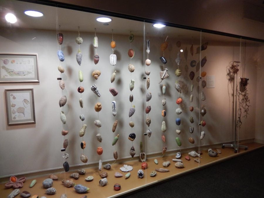 A glass display box filled with 10 hanging garlands of small multicolored, circular sculptures. Other sculptures are placed on the floor around the hanging display.