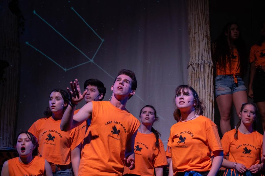 A crowd of actors in orange shirts look up, with a constellation projected behind them.