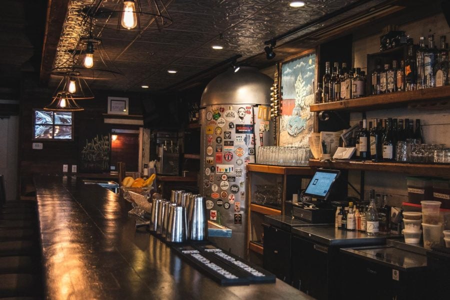 A+bar+is+lit+by+vintage+lights.+There+are+shelves+with+bottles+of+liquor.
