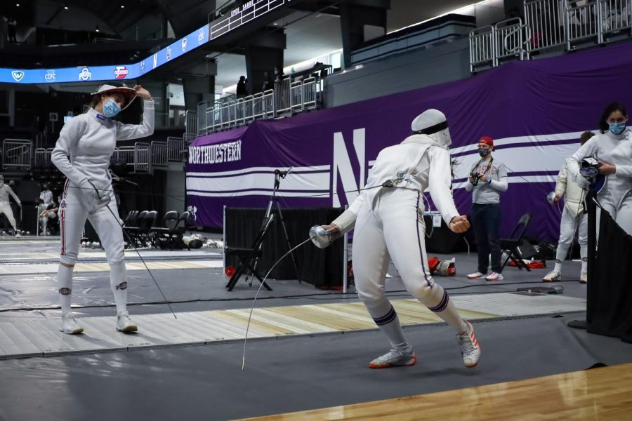 The fencer on the right leans back in rejoice after a match with their opponent on the left, who is taking off their head attire.
