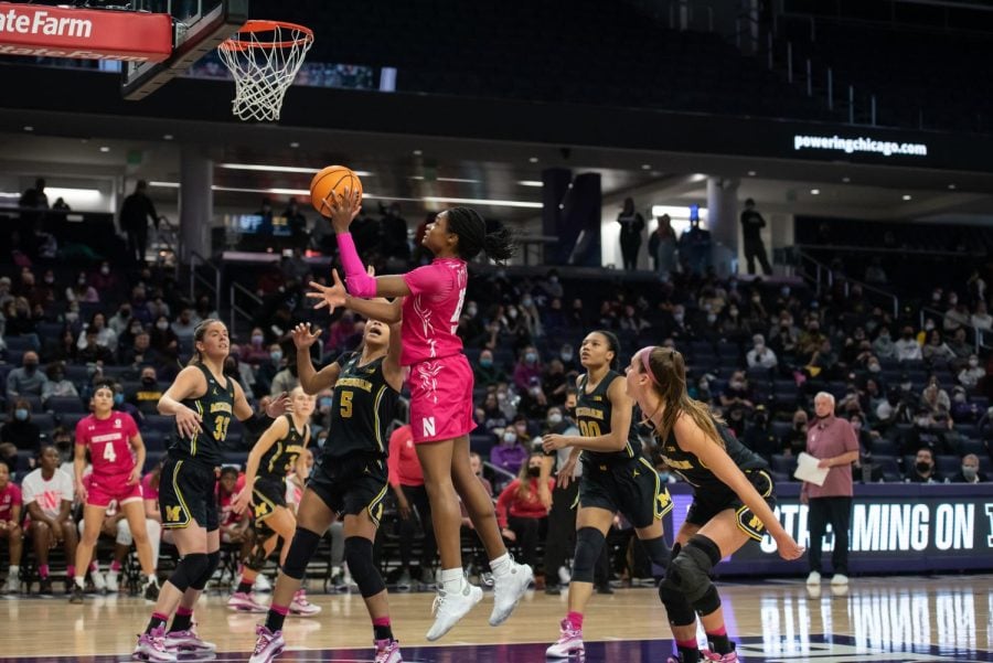 A person in a pink jersey surrounded by people in black jerseys jumps and pushes a basketball towards a hoop.